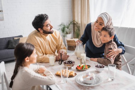 Foto de Smiling african american woman in hijab hugging son near family during suhur breakfast in morning - Imagen libre de derechos