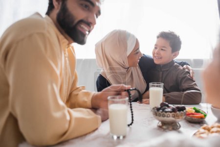 Téléchargez les photos : Mère afro-américaine positive dans le hijab étreignant son fils près de la famille et suhur petit déjeuner à la maison - en image libre de droit