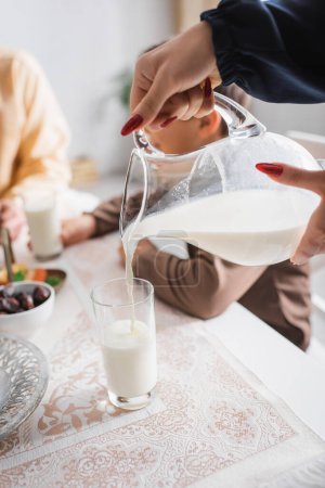 Téléchargez les photos : Vue recadrée de la femme musulmane versant du lait dans du verre près de la famille floue le matin - en image libre de droit