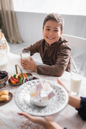 Foto de Cheerful muslim boy holding milk and looking at mom holding turkish delight during ramadan - Imagen libre de derechos