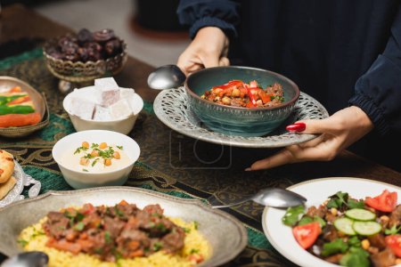 Foto de Vista recortada de la mujer musulmana sosteniendo deliciosa comida cerca de la mesa durante la cena de ramadán - Imagen libre de derechos