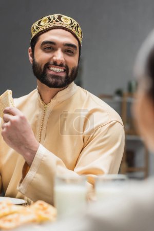 Photo for Smiling muslim father holding pita bread near blurred daughter at home - Royalty Free Image