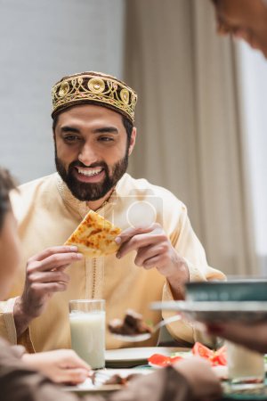 Uśmiechnięty muzułmański ojciec trzymający chleb pita w pobliżu rozmytej rodziny podczas kolacji Ramadan 