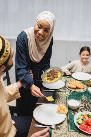 Téléchargez les photos : Femme afro-américaine en hijab servant pilaf dans une assiette près du mari pendant le ramadan à la maison - en image libre de droit