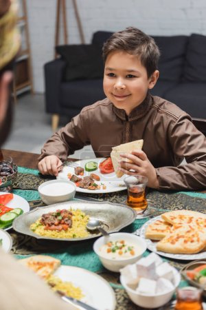 Téléchargez les photos : Garçon musulman souriant tenant du pain pita près du père flou et de la nourriture à la maison - en image libre de droit