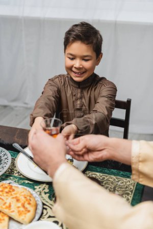 Téléchargez les photos : Souriant garçon musulman donnant verre de thé turc à papa flou pendant iftar à la maison - en image libre de droit