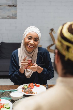 Téléchargez les photos : Femme afro-américaine souriante dans le hijab tenant le thé près du mari flou pendant iftar à la maison - en image libre de droit
