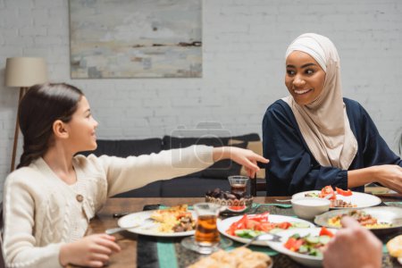 Téléchargez les photos : Mère afro-américaine souriante dans le hijab regardant sa fille près de la nourriture iftar à la maison - en image libre de droit