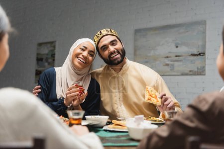 Téléchargez les photos : Homme musulman souriant étreignant sa femme avec du thé près des enfants flous et de la nourriture à la maison - en image libre de droit