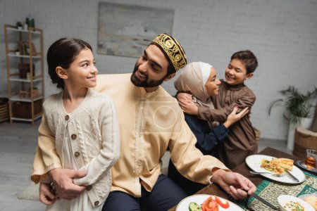 Foto de Muslim father hugging daughter near family and ramadan dinner at home - Imagen libre de derechos
