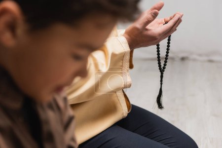Téléchargez les photos : Homme musulman avec des perles de prière priant près de son fils flou à la maison - en image libre de droit