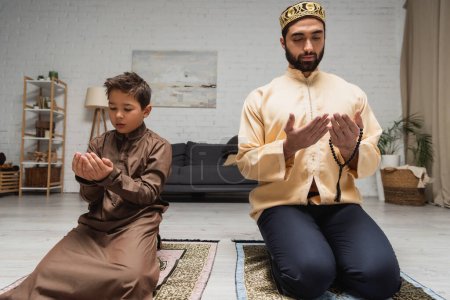 Foto de Hombre musulmán e hijo rezando en alfombras durante el ramadán en casa - Imagen libre de derechos
