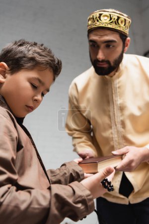 Zamazany muzułmański ojciec daje księgę synowi z kulkami modlitewnymi w domu 