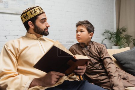 Photo for Muslim father holding book and looking at son on couch at home - Royalty Free Image