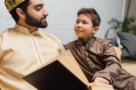 Photo pour Garçon musulman regardant papa près du livre flou à la maison - image libre de droit