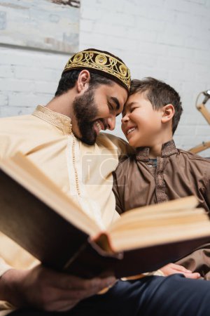 Photo for Cheerful middle eastern man and son reading blurred book at home - Royalty Free Image
