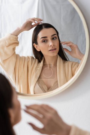 young brunette woman adjusting hair and looking at mirror in bathroom 
