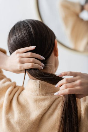 Téléchargez les photos : Vue arrière de la jeune femme brune ajuster les cheveux près du miroir dans la salle de bain - en image libre de droit