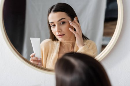 brunette young woman in bathrobe holding tube with cream and looking at mirror in bathroom 
