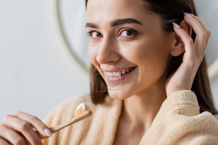 portrait of cheerful woman in bathrobe holding toothbrush with toothpaste while adjusting hair 