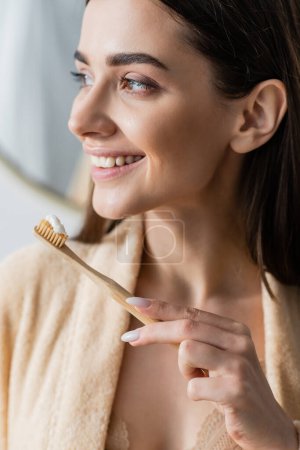 Foto de Mujer joven sonriente en albornoz sosteniendo cepillo de dientes con pasta de dientes en el baño - Imagen libre de derechos