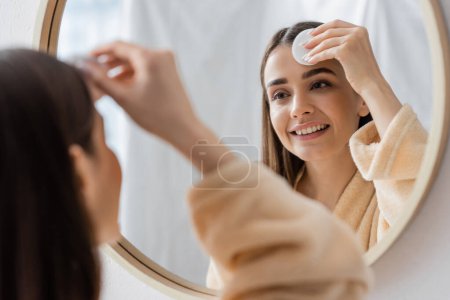 Photo for Reflection of smiling young woman cleansing face with cotton pad in bathroom mirror - Royalty Free Image
