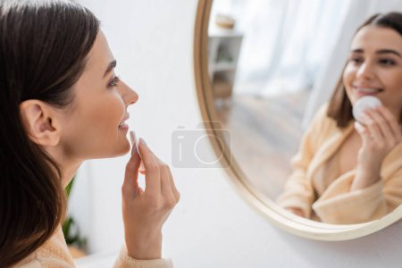 uśmiechnięta młoda kobieta w szlafroku oczyszczająca twarz z podkładką bawełnianą i patrząca w lustro w łazience 