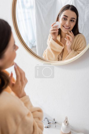 reflection of pleased young woman cleansing face with cotton pad in bathroom mirror 