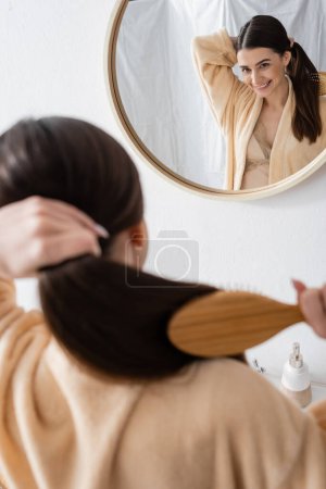 reflection of happy young woman brushing shiny hair in bathroom mirror 