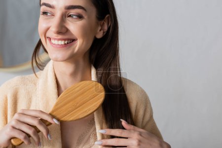 joyful young woman in bathrobe brushing shiny hair and looking away in bathroom 