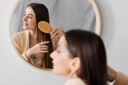 reflection of cheerful woman brushing shiny hair near mirror in bathroom 