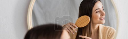 reflection of cheerful woman brushing shiny hair near mirror in bathroom, banner 