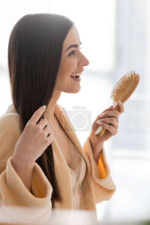 Photo for Side view of happy young woman holding wooden hair brush in bathroom - Royalty Free Image