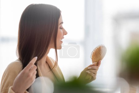 Photo for Side view of cheerful young woman holding wooden hair brush in bathroom - Royalty Free Image
