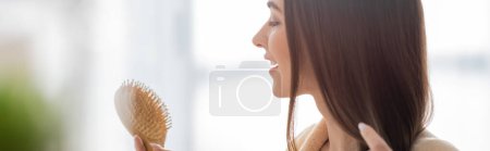 Photo for Side view of cheerful young woman holding wooden hair brush in bathroom, banner - Royalty Free Image