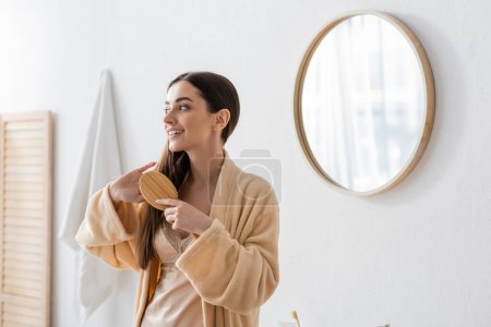 happy young woman in bathrobe brushing hair in white modern bathroom 