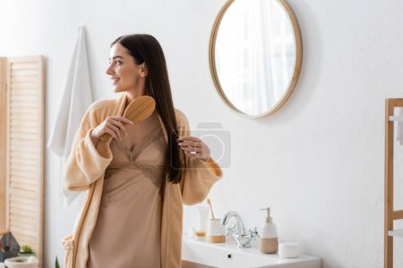 cheerful young woman in bathrobe brushing hair in white modern bathroom 