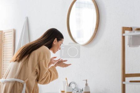 junge brünette Frau mit geschlossenen Augen wäscht Gesicht im Badezimmer 