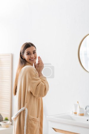 positive woman in bathrobe looking at camera near mirror in bathroom 