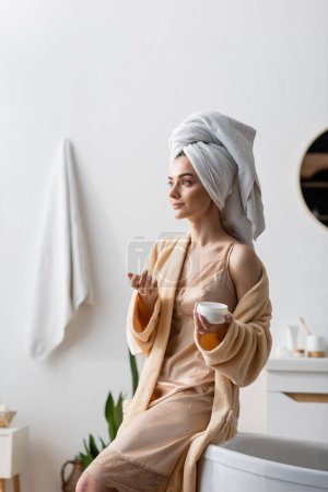 Foto de Young woman with towel on head holding container with body butter in bathroom - Imagen libre de derechos