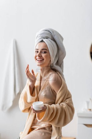 Foto de Joyful young woman with towel on head and bathrobe holding container with body butter in bathroom - Imagen libre de derechos
