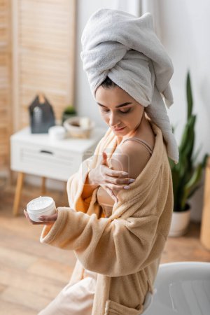 young woman in bathrobe with towel on head holding container while applying body butter 