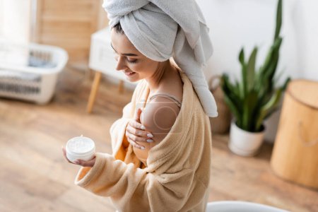 smiling woman in bathrobe with towel on head holding container with body butter 