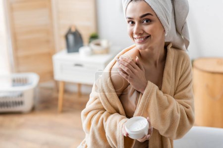Photo for Happy young woman in bathrobe with towel on head holding container while applying body butter - Royalty Free Image