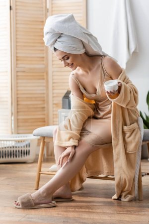 Photo for Cheerful young woman in bathrobe and slippers applying cream on leg in bathroom - Royalty Free Image