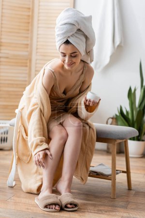 Cheerful woman in nightgown and bathrobe applying cream on leg in morning
