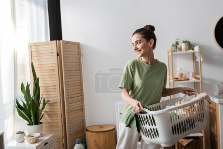 Cheerful young woman holding basket with clothes in laundry room 