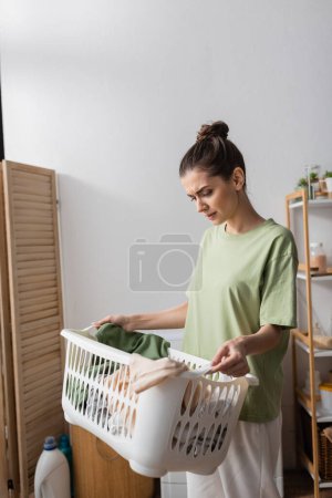 Unzufriedene Frau blickt auf Korb mit Kleidung in Waschküche 