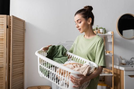 Foto de Young brunette woman holding basket with clothes in laundry room - Imagen libre de derechos