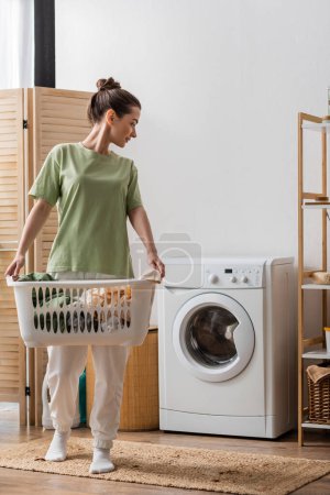 Photo for Side view of young woman holding basket with clothes and looking at washing machine at home - Royalty Free Image